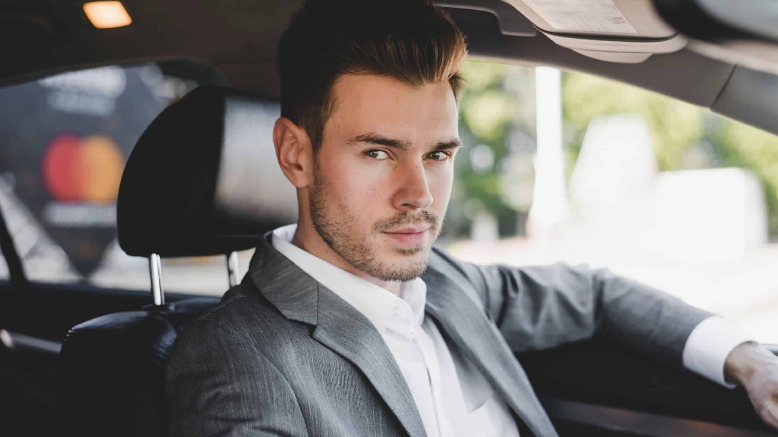 An attractive man wearing a sleek suit sitting in a car.