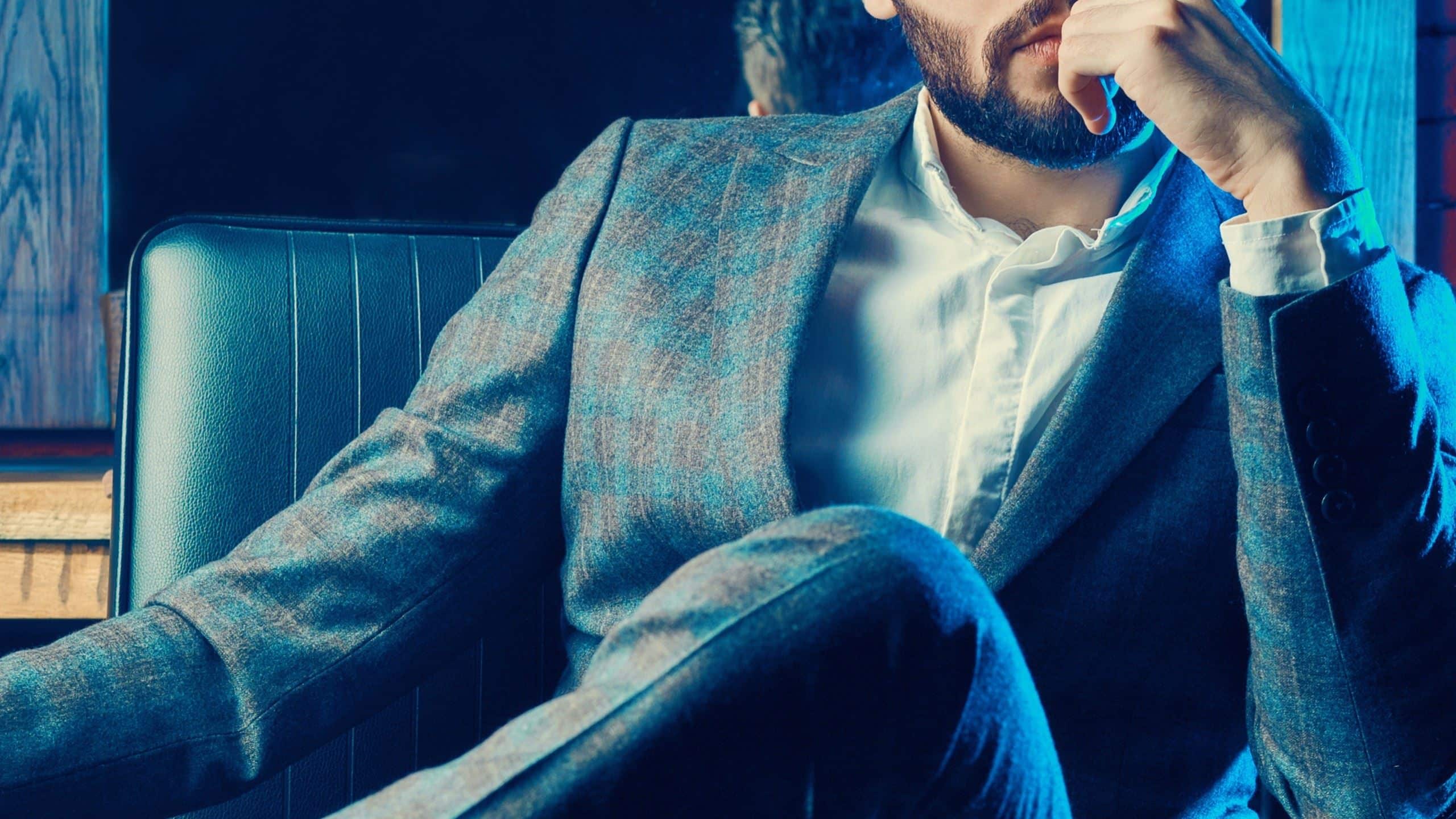 A man in a fashionable suit sitting in a chair in a confident pose.